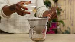 A pair of hands pouring oils through a strainer and into a glass measuring cup from Arina Soler's Make Herbal Tinctures Creativebug class.