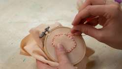 A pair of women's hands holding a small embroidery hoop and stitching on a piece of beige fabric with bright red thread.