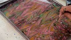 A tray of size with purple, black, and green marbling ink dropped on top and a hand moving a marbling comb through the inks. 