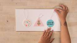 Three ornaments, one white in the shape of a circle, one pink and triangular with a Christmas tree, one teal with decorative elements, on a table