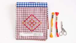 A sample page from Blair Stocker's Chicken Scratch Embroidery Sampler Book class on Creativebug on mauve gingham plus two skeins of DMC thread in yellow and red and stork-shaped thread snips.