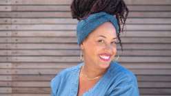 A woman in a blue shirt with pink lipstick and gold jewelry wearing a twist rib knit headband from Faith Hale's Creativebug Knit Headbands Three Ways class.